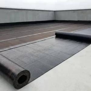 Bitumineuze dakbedekking die wordt uitgerold op een plat dak, met overlappende naden en betonnen dakrand op de achtergrond.