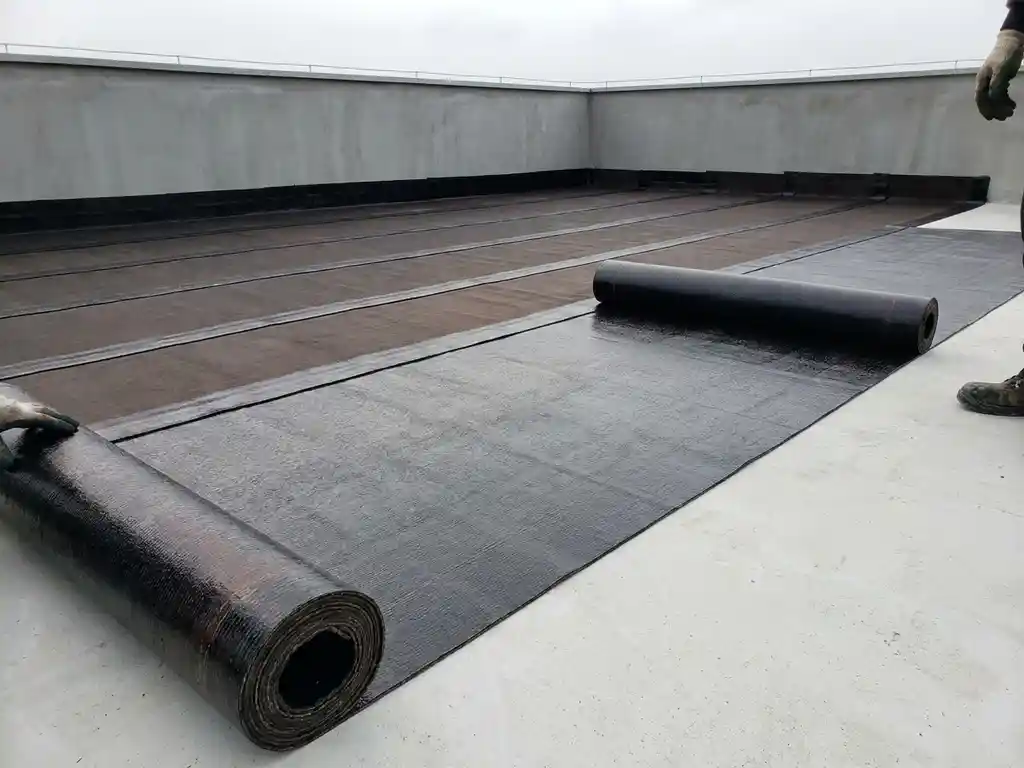 Bitumineuze dakbedekking die wordt uitgerold op een plat dak, met overlappende naden en betonnen dakrand op de achtergrond.