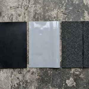 Drie dakbedekkingsmonsters naast elkaar op betonnen plat dak: mat zwart EPDM, glanzend grijs PVC en getextureerd APP bitumenfolie.