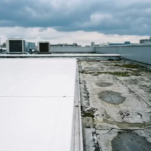 Industrieel plat dak met nieuw wit membraan naast verouderd grijs bitumen dakbedekking, bewolkte Nederlandse lucht.