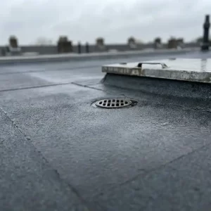 Plat dak met bitumineuze dakbedekking, afvoerput en inspectieluik onder bewolkte Nederlandse lucht.