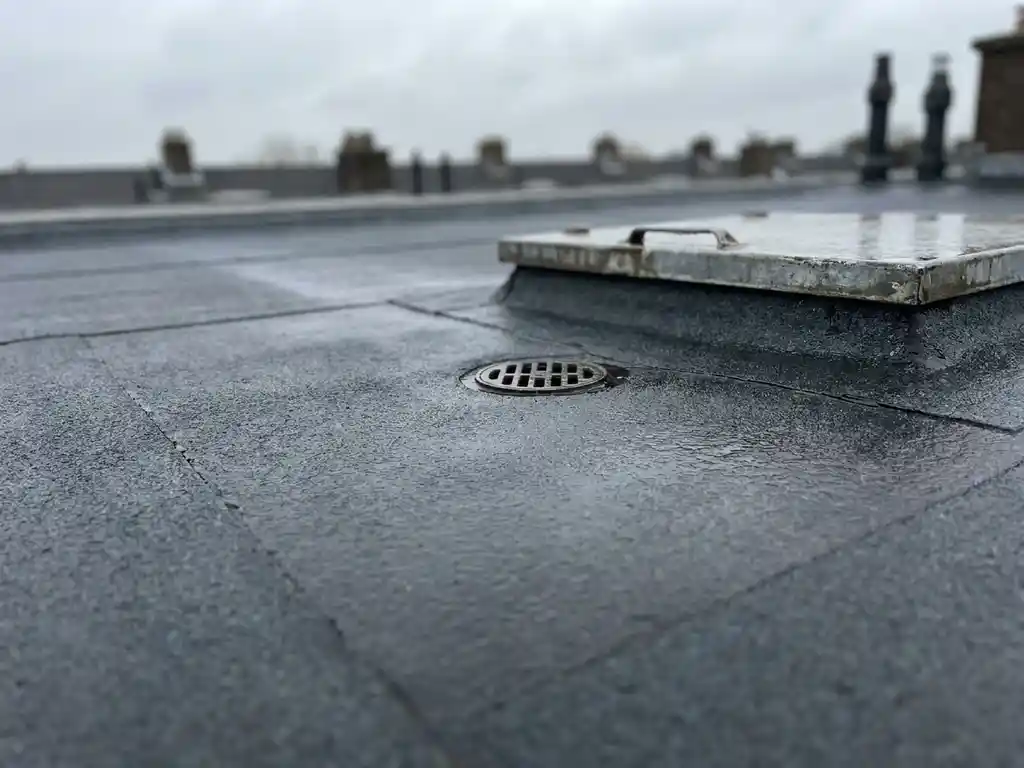 Plat dak met bitumineuze dakbedekking, afvoerput en inspectieluik onder bewolkte Nederlandse lucht.