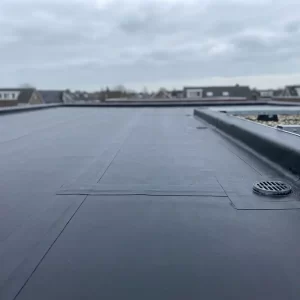 Vers aangebrachte bitumenmembraan op plat dak met afwateringspunt, donker asfaltoppervlak onder bewolkte Nederlandse lucht.
