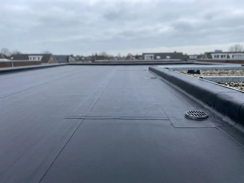 Vers aangebrachte bitumenmembraan op plat dak met afwateringspunt, donker asfaltoppervlak onder bewolkte Nederlandse lucht.