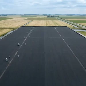 Bovenaanzicht van een groot commercieel plat dak met donkere bitumenmembraan, omgeven door een Nederlands polderlandschap onder bewolkte lucht.