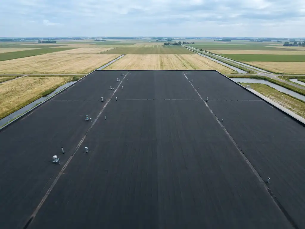 Bovenaanzicht van een groot commercieel plat dak met donkere bitumenmembraan, omgeven door een Nederlands polderlandschap onder bewolkte lucht.