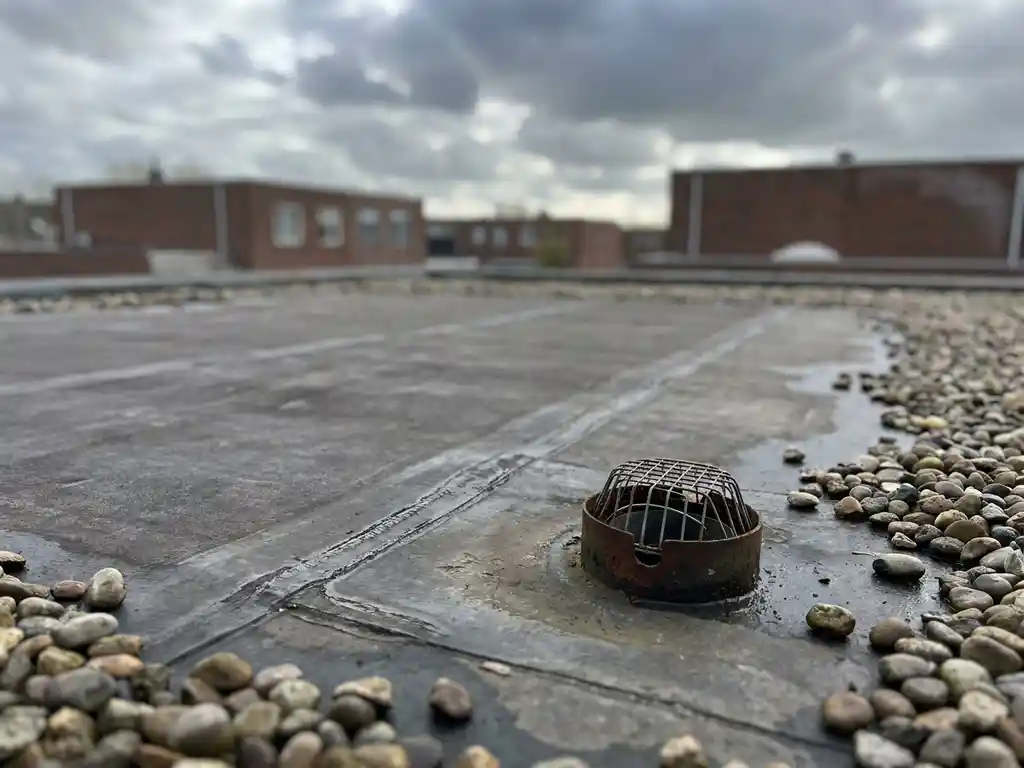 Verweerd EPDM-dakbedekking op een plat commercieel dak in Nederland, met naadlijnen, afvoerput en grindrand onder bewolkte lucht.