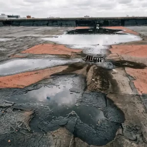 Verweerd plat dak met staand water, gebarsten bitumenmembraan en vochtsporen bij afvoerput onder bewolkte lucht.