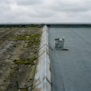 Plat dak half bedekt met verouderde gebarsten bitumen en mos, half met nieuw dakbedekkingsfolie, gereedschap en kit op de grens.