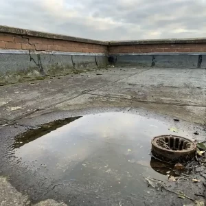 Verweerd plat dak met zichtbare membraannaden, waterplassen bij verstopte afvoer en gebarsten bitumen langs de dakrand.
