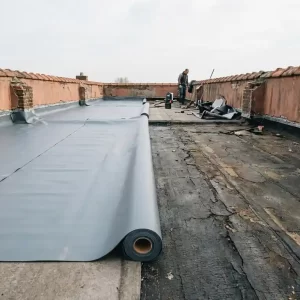 Plat dak in renovatie met deels uitgerolde grijze EPDM-membrane naast verouderd gebarsten bitumen dakbedekking, lichtbewolkte lucht op achtergrond.