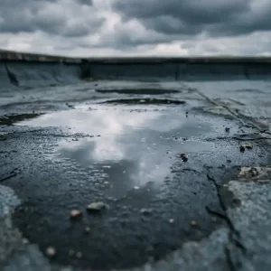Stilstaand water in een lage plek op een bitumen platdak, met bewolkte grijze lucht weerspiegeld in de plas.